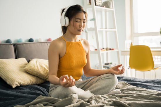 Relaxed young calm woman wearing headphones sitting at home in the bed in lotus position, while meditating and practicing yoga with pleasure smile and closed eyes. Enjoying of the morning concept